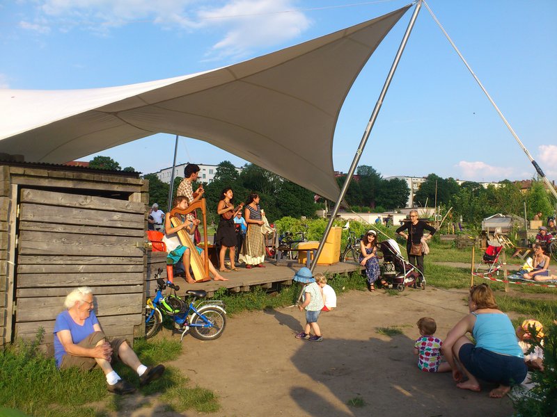 Menschen auf einer kleinen Bühne unter einem Sonnensegel musizieren gemeinsam im Sommer